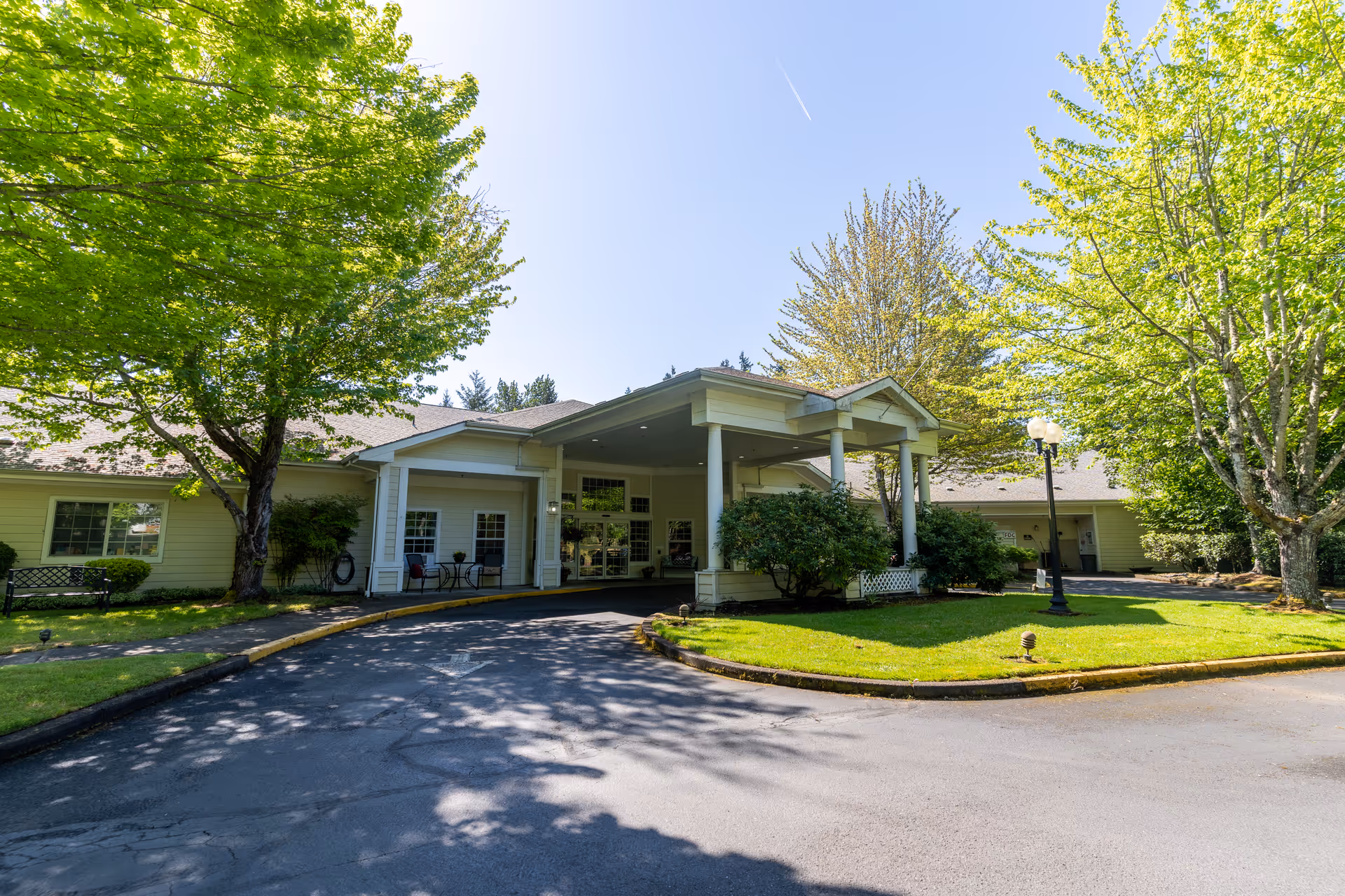 Exterior view of Meadow Creek Village Assisted Living facility entrance with a covered drop-off area, surrounded by green trees and a well-maintained lawn under a clear blue sky.