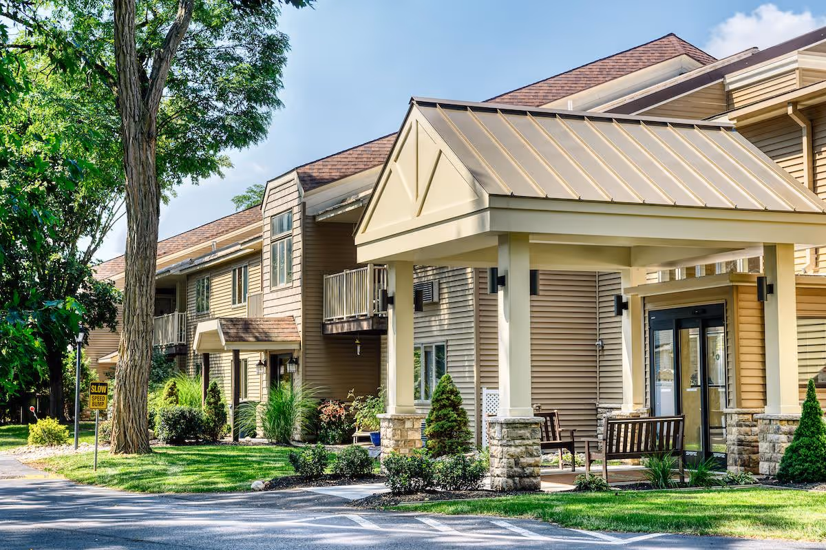 Exterior view of Kinsington Oak Grove facility showing a covered entrance with stone pillars, beige siding, and a well-maintained lawn with trees and shrubs under a clear sky.