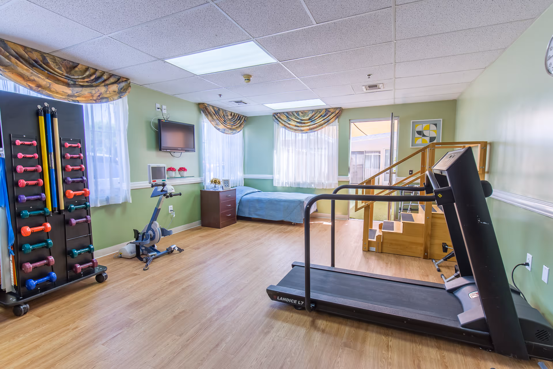 A bright room with light green walls and wood flooring featuring exercise equipment including a treadmill, stationary bike, and a rack of colorful dumbbells. There is a single bed with a blue blanket near the windows, a small wooden dresser, a wall-mounted TV, and a set of wooden stairs with handrails leading to a platform. The room has large windows with sheer white curtains and patterned valances, and a clock is visible on the wall.
