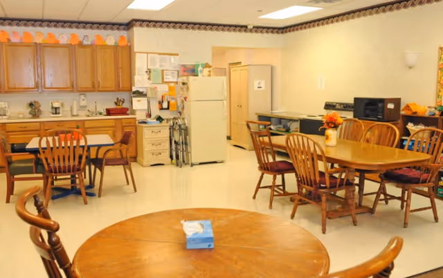A communal dining and kitchen area with wooden tables and chairs. The kitchen area has wooden cabinets, a refrigerator, a microwave, and various kitchen appliances. The room is decorated with a border of small orange pumpkin decorations along the top of the cabinets and a vase with flowers on one of the tables.