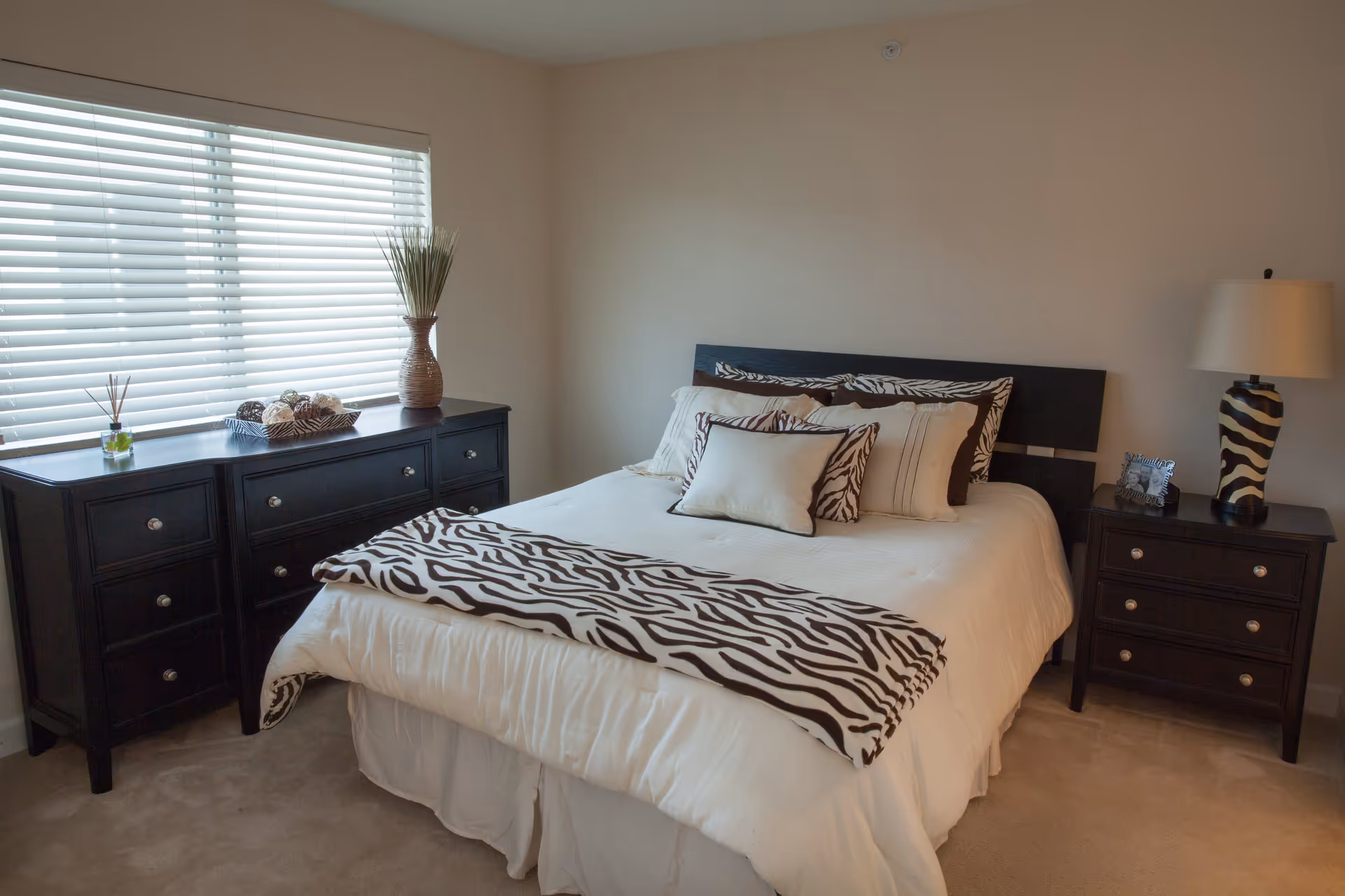 A bedroom with a neatly made bed featuring white bedding and zebra-patterned pillows and throw. There is a dark wooden dresser with decorative items and a vase with tall grass next to a window with blinds. A matching dark wooden nightstand with a zebra-patterned lamp and a small framed photo is beside the bed.