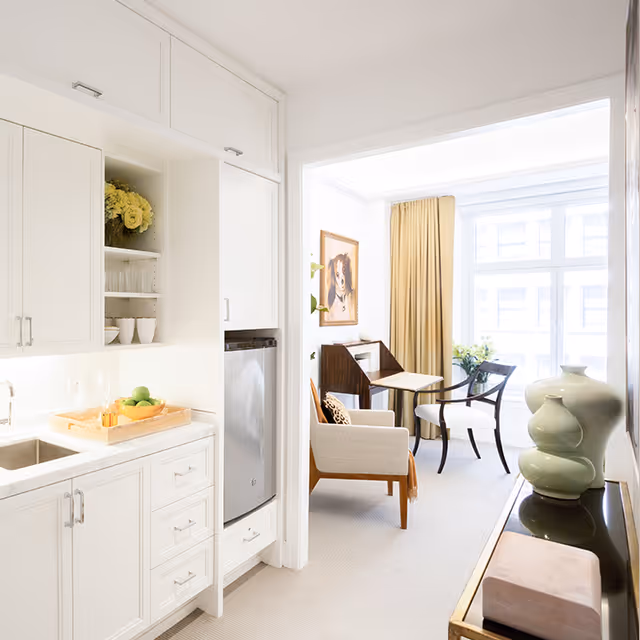 Bright and airy interior space featuring a small kitchen area with white cabinets, a sink, and a countertop with a tray holding green apples and glasses. Adjacent to the kitchen is a cozy sitting area with a beige armchair, a small table, two chairs, a framed dog portrait on the wall, and large windows with beige curtains letting in natural light. A decorative green vase sits on a console table near the entrance to the sitting area.