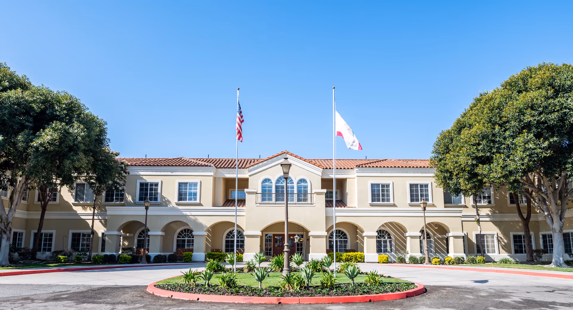 Front exterior view of San Juan Hills Healthcare Center building with two flagpoles displaying the American flag and another flag, surrounded by trees and landscaping under a clear blue sky.