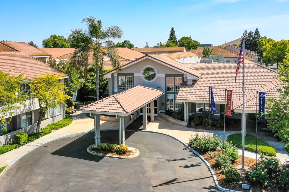 Exterior view of a senior living facility with a covered entrance, surrounded by trees and landscaping. The building has a tiled roof and multiple windows, with flags and a flagpole near the entrance.