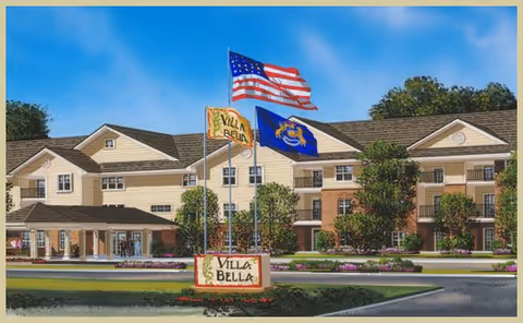Exterior view of Villa Bella senior living facility with a large multi-story building, landscaped grounds, and three flags flying on flagpoles including the American flag, a Villa Bella flag, and a state flag.