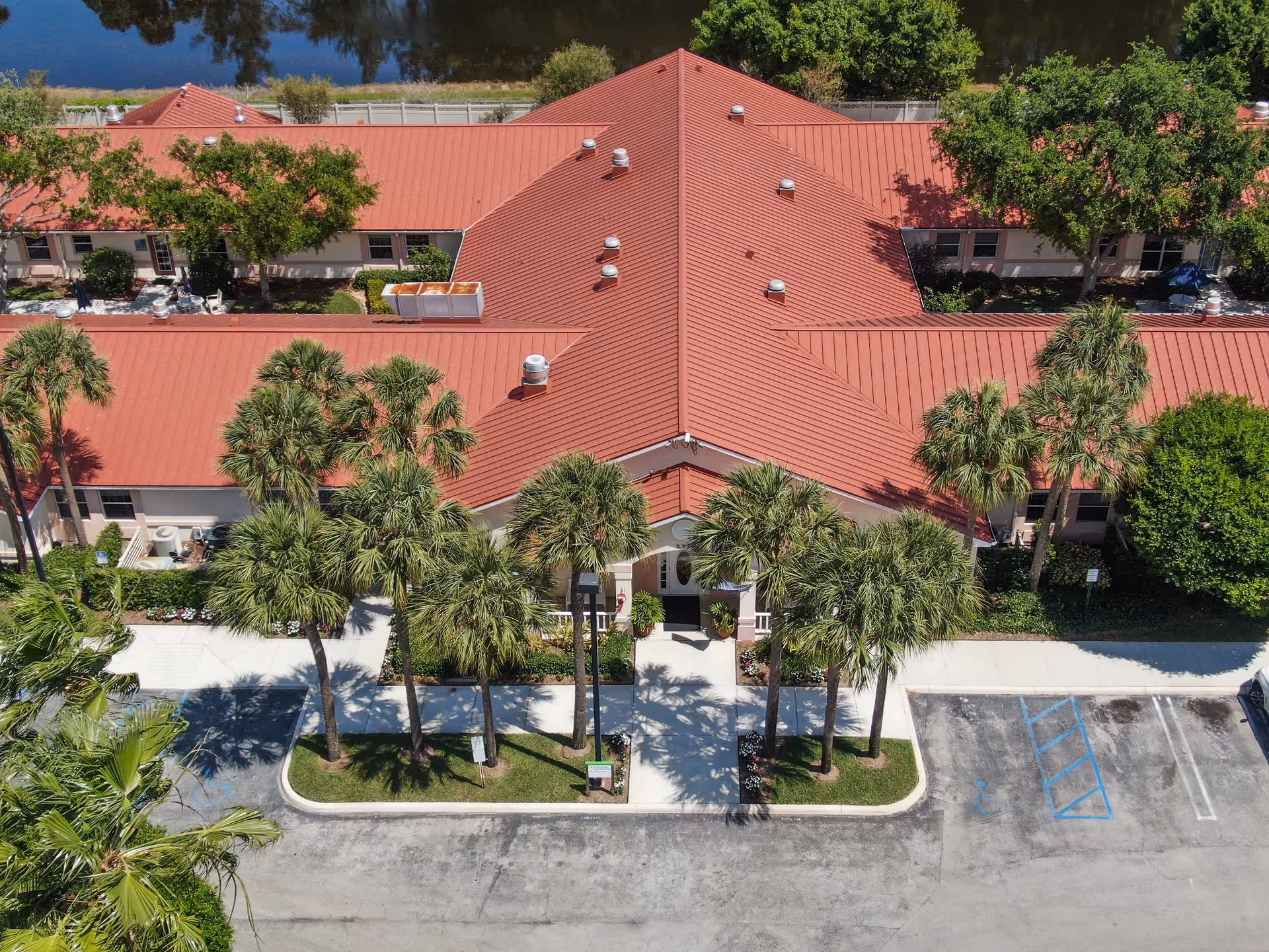 Aerial view of a single-story building with a red roof surrounded by palm trees and greenery. The building has a central entrance with a walkway leading to a parking area with marked handicap spaces. The surrounding area includes trees and a body of water in the background.