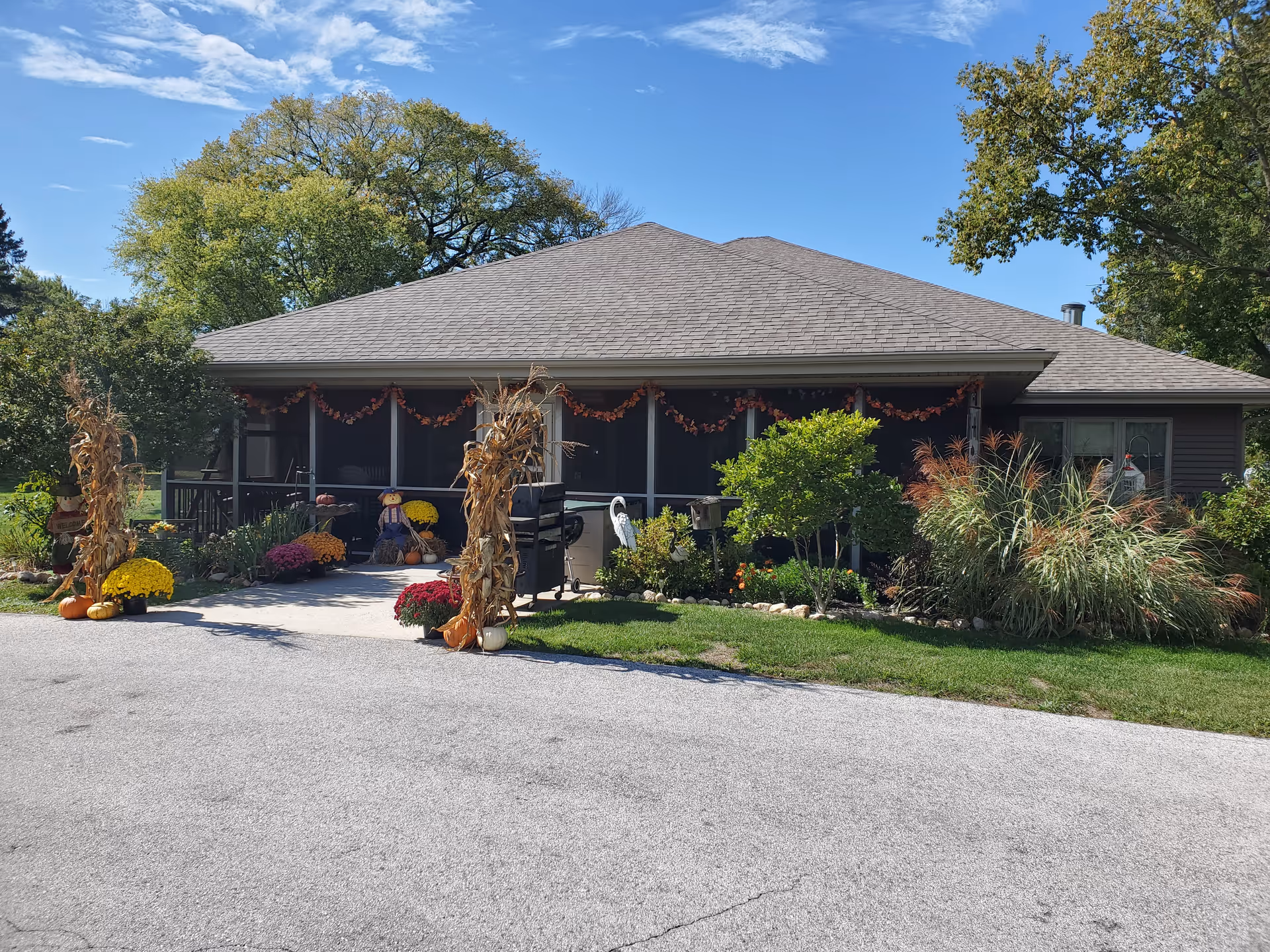 Exterior view of a single-story building with a gray roof and screened porch decorated with autumn-themed garlands. The entrance pathway is flanked by dried corn stalks, pumpkins, and colorful potted flowers. Surrounding the building are green trees, shrubs, and a well-maintained lawn under a blue sky with scattered clouds.