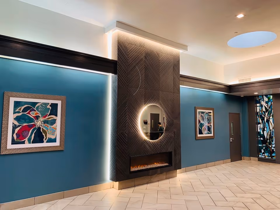 Interior view of a modern room with teal walls, two colorful framed artworks, a dark textured fireplace with a circular mirror above it, and a decorative stained glass panel near a closed door. The ceiling has recessed lighting and a circular skylight.