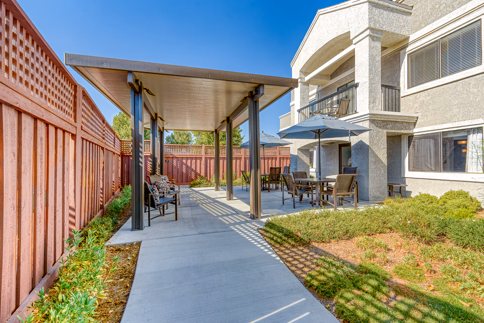Outdoor patio area at Menifee Senior Living featuring a covered walkway with benches and a seating area with tables, chairs, and umbrellas next to a beige stucco building under a clear blue sky.
