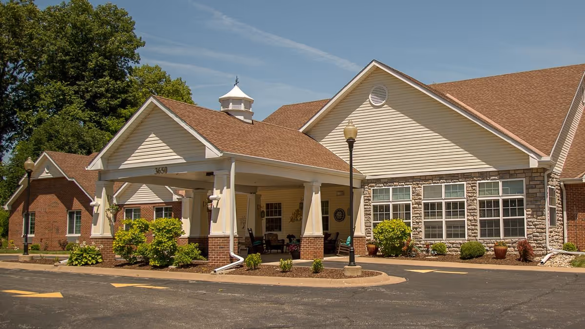 Exterior view of a senior living facility building with a covered entrance supported by white columns. The building has beige siding, stone accents, and a brown shingled roof. There are shrubs and small plants around the building and a clear blue sky above.