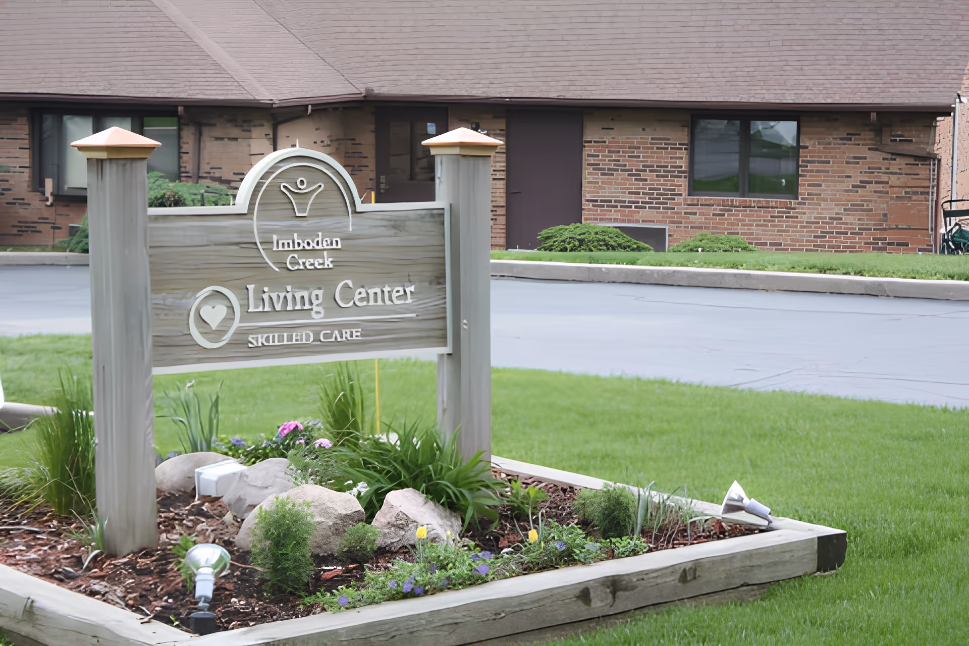 A wooden sign for Imboden Creek Living Center Skilled Care is displayed in a landscaped garden bed with rocks, flowers, and greenery. Behind the sign is a brick building with windows and a door, and a paved driveway.