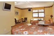 A cozy bedroom in a senior living facility with two beds covered in floral patterned bedspreads. The room has a wall-mounted television, two chairs with a small table between them, a window with a valance, and a few decorative items including a framed picture and a vase with flowers.