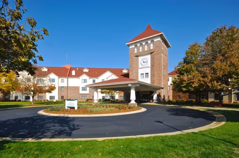 Exterior view of a senior living facility building with a prominent clock tower, red roof, and brick facade. The building is surrounded by green grass, trees with autumn foliage, and a circular driveway with a covered entrance.