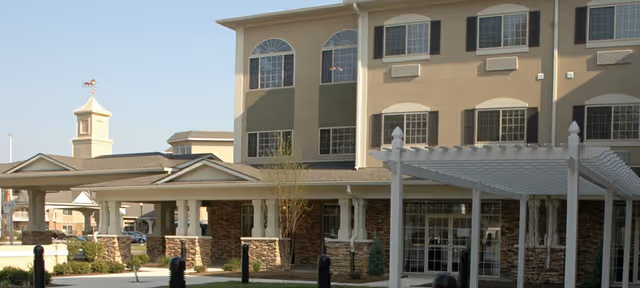 Exterior view of a multi-story senior living facility building with large windows, a covered entrance supported by stone pillars, and a white pergola structure in front. The sky is clear and the landscaping includes small trees and shrubs.