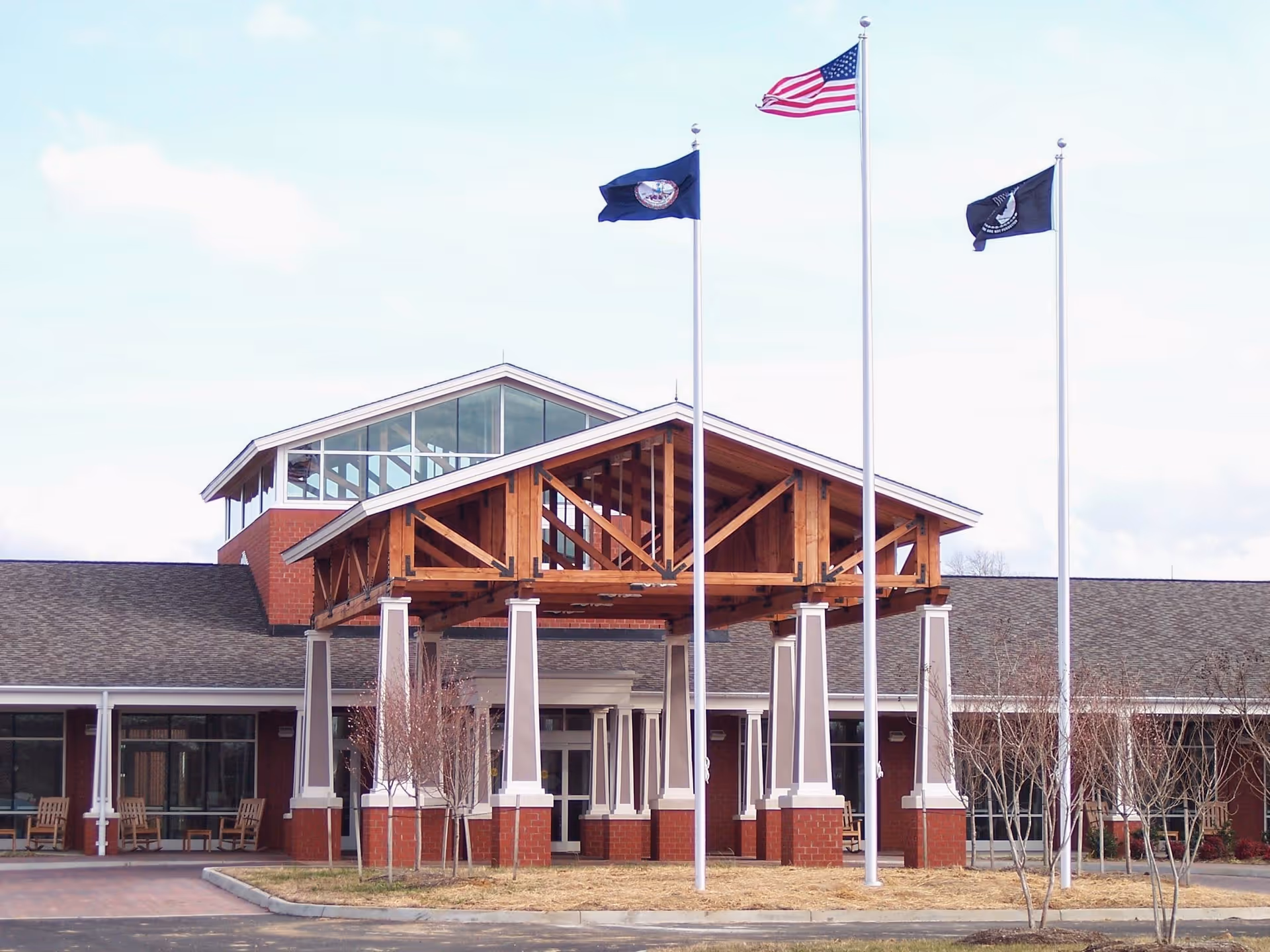 Front exterior view of the Commonwealth of Virginia: Sitter & Barfoot Veterans Care Center building with a covered entrance supported by columns, three flagpoles flying the American flag and two other flags, and some leafless trees in front.