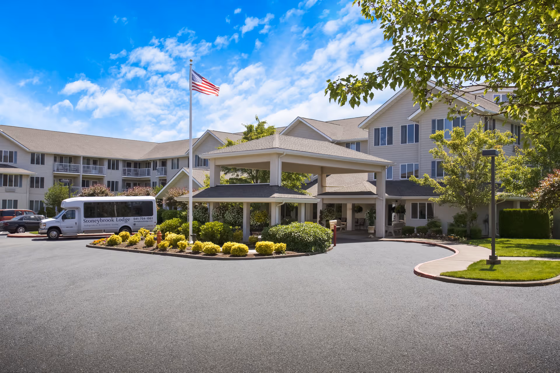 Exterior view of Stoneybrook Lodge senior living facility with a covered entrance, landscaped garden area, an American flag on a flagpole, and a shuttle van parked nearby under a partly cloudy blue sky.