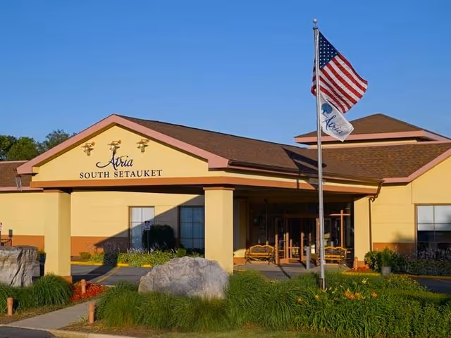 Exterior front view of Atria South Setauket senior living community building with a flagpole displaying the American flag and a white flag with the Atria logo, surrounded by greenery and rocks under a clear blue sky.