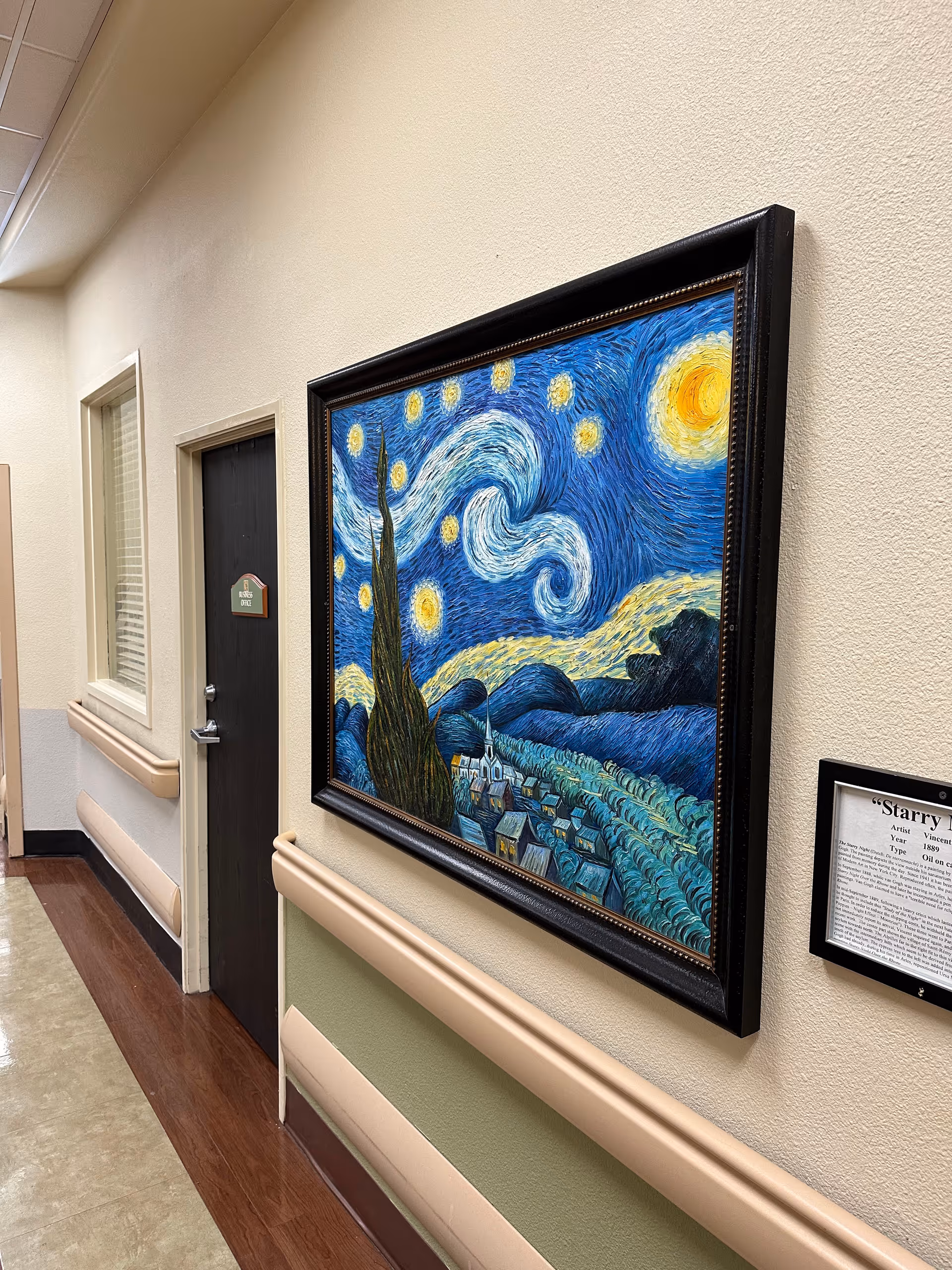 A hallway in a nursing and rehabilitation center with beige walls and wooden floor trim. On the right wall, there is a framed reproduction of Vincent van Gogh's painting 'Starry Night' along with a small informational plaque. On the left side, there is a door labeled 'Office 002' and a window with blinds.
