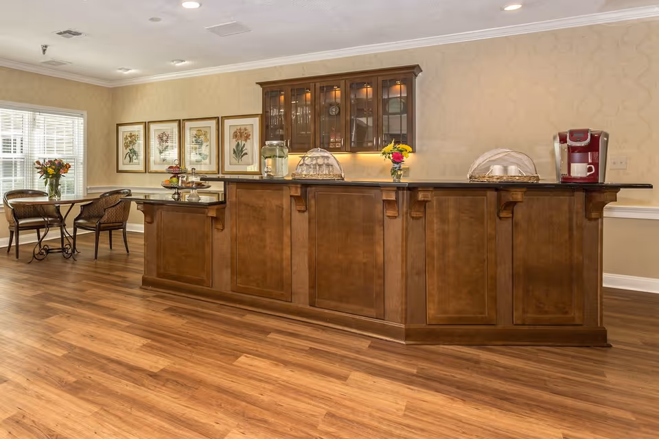 A warm and inviting interior space featuring a long wooden counter with a black countertop. On the counter are a glass jar with water and lemon slices, a tiered tray with pastries, a covered dish, a vase with flowers, and a red coffee maker with a white cup. Behind the counter is a wooden cabinet with glass doors displaying glassware. To the left, there is a small dining area with a round glass table, two chairs, a vase with flowers, and four framed botanical prints on the wall. The room has wood flooring and beige patterned wallpaper.