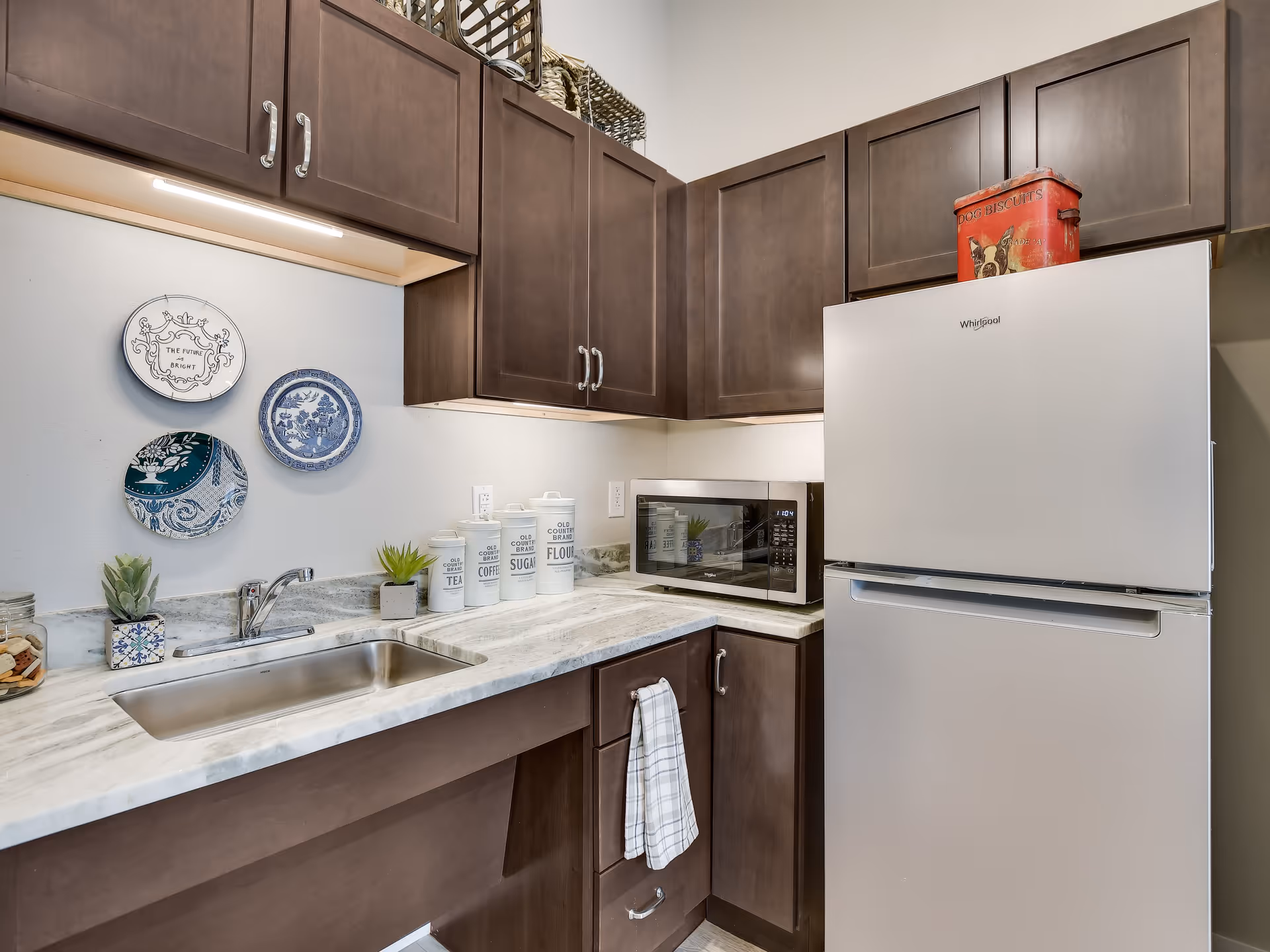 A compact kitchen area with dark brown cabinets, a stainless steel sink, a white Whirlpool refrigerator with a red dog biscuits tin on top, a microwave, and decorative plates on the wall. The countertop has canisters labeled tea, coffee, sugar, and flour, along with small potted plants.