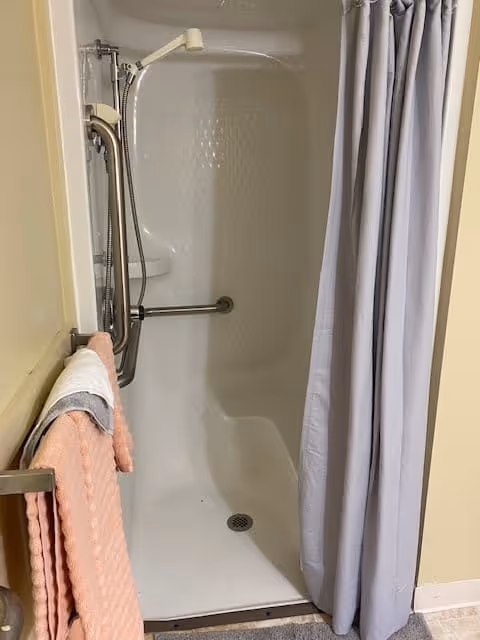 A small shower stall with a white curtain partially drawn to the side. Inside the shower, there is a handheld showerhead attached to a vertical grab bar, and a horizontal grab bar is mounted on the wall. A peach-colored towel and a white towel hang on a towel rack outside the shower.