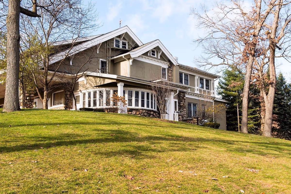 A large two-story house with beige exterior walls and stone accents on the pillars and lower walls, surrounded by leafless trees and green grass on a sunny day with a blue sky and some clouds.