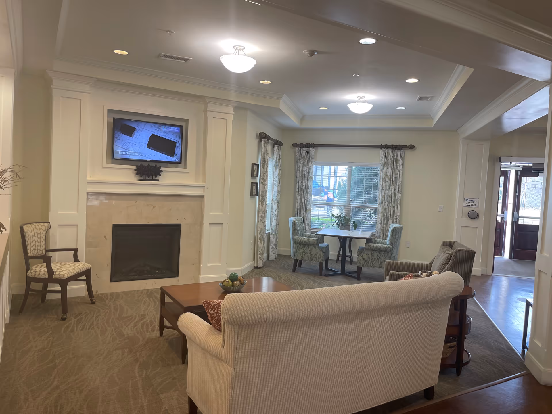 A cozy living room area in Carriage Inn Katy featuring a beige sofa, two armchairs, a wooden coffee table, a fireplace with a mounted TV above it, and a small dining table with four chairs near a window with floral curtains.