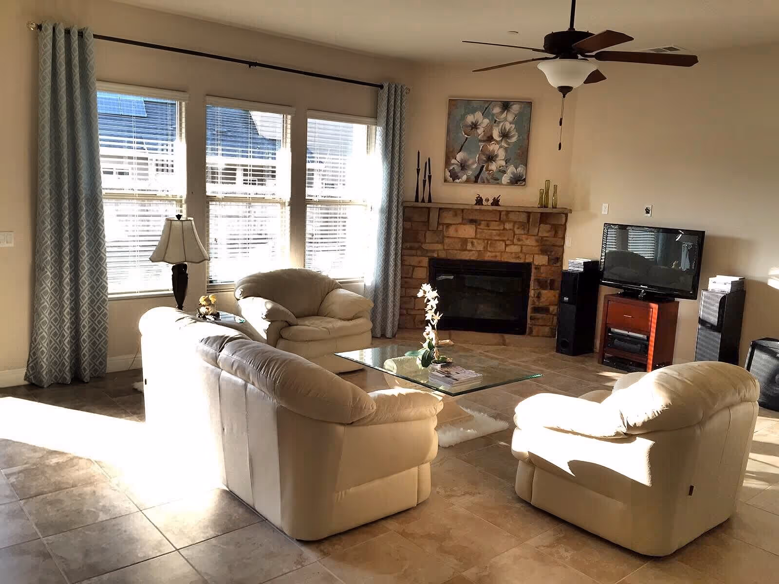 A bright living room with beige leather sofas and armchairs arranged around a glass coffee table. The room features a stone fireplace with decorative items and a floral painting above it. There is a flat-screen TV on a wooden stand with speakers on either side. Large windows with patterned curtains let in natural light, and a ceiling fan hangs above.