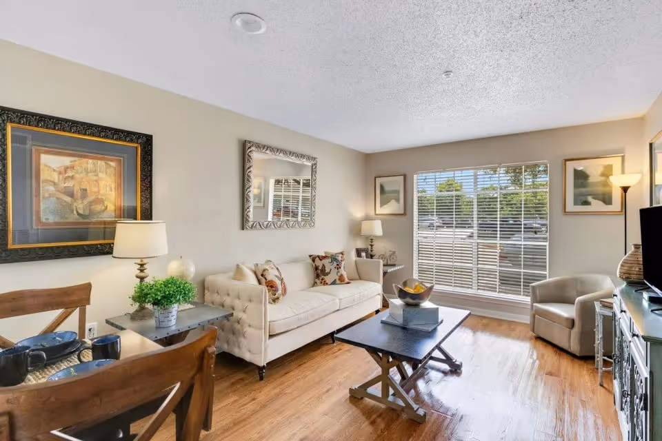 A bright living room with a beige tufted sofa adorned with floral pillows, a wooden coffee table with decorative items, a beige armchair, and a TV on a stand. The room has wooden flooring, a large window with white blinds letting in natural light, framed artwork on the walls, and two lamps providing additional lighting.