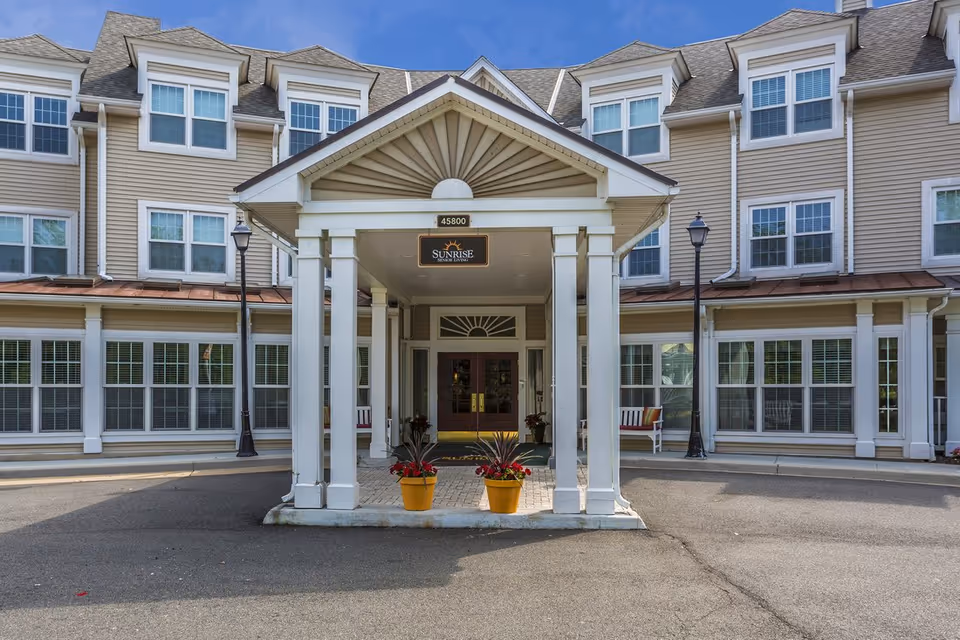 Front entrance of a multi-story senior living facility with a covered driveway, two potted plants with red flowers, and a sign above the entrance that reads 'Sunrise Senior Living'. The building has multiple windows and beige siding with white trim.