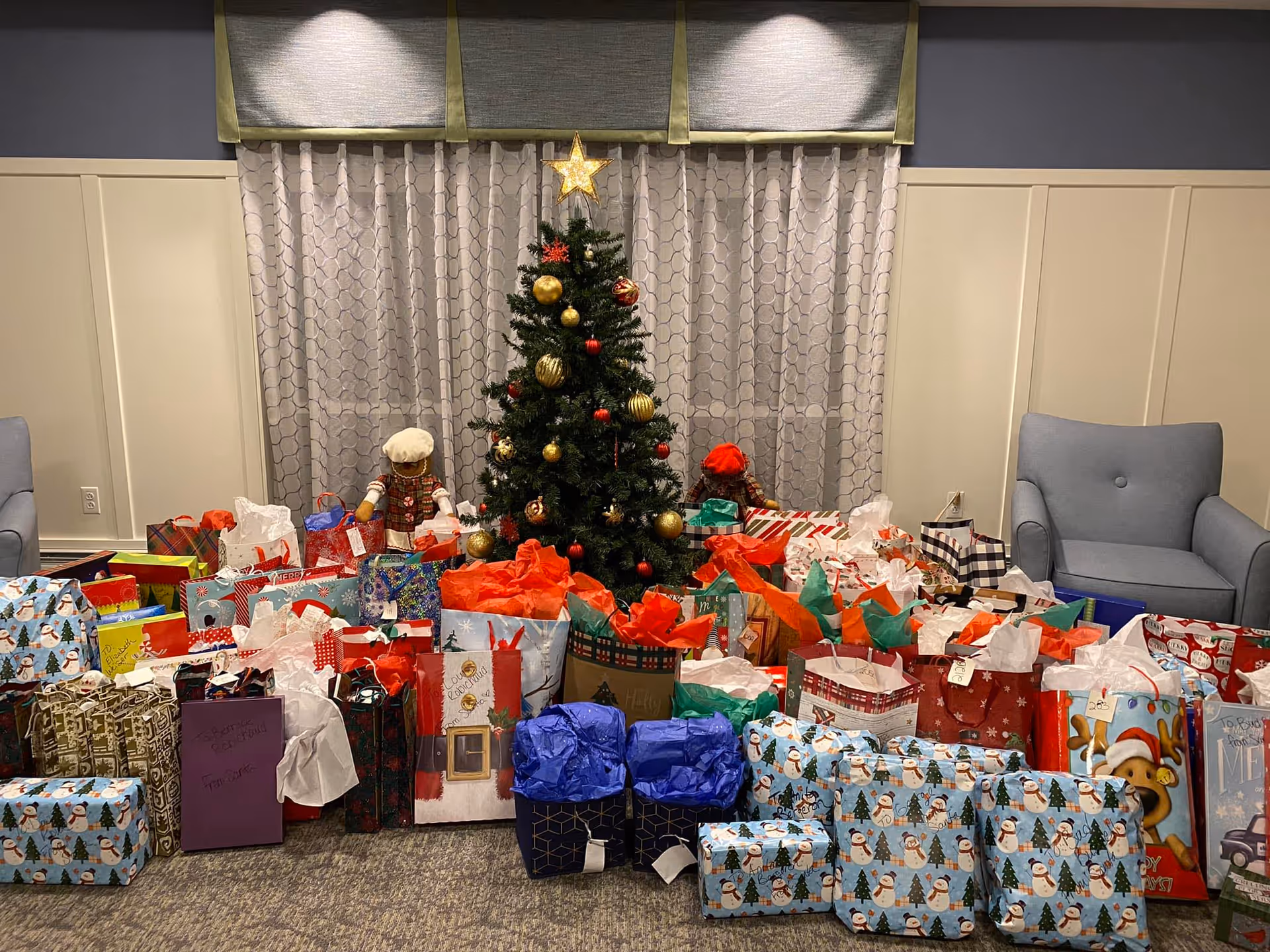 A decorated Christmas tree with a glowing star on top is centered in a room with two armchairs on either side. The floor in front of the tree is filled with numerous wrapped holiday gifts and gift bags in various colors and patterns. Behind the tree is a window with patterned curtains and a valance. Two dolls dressed in holiday attire are placed behind the gifts on either side of the tree.