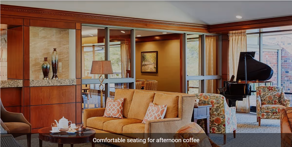 A cozy living room area with a beige sofa adorned with patterned cushions, floral armchairs, a round wooden coffee table set with a teapot and cups, a floor lamp, and a grand piano near large windows letting in natural light.