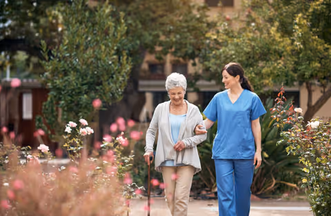 An elderly woman with a walking cane is being assisted by a female healthcare worker in blue scrubs as they walk together along a garden path surrounded by greenery and flowers.