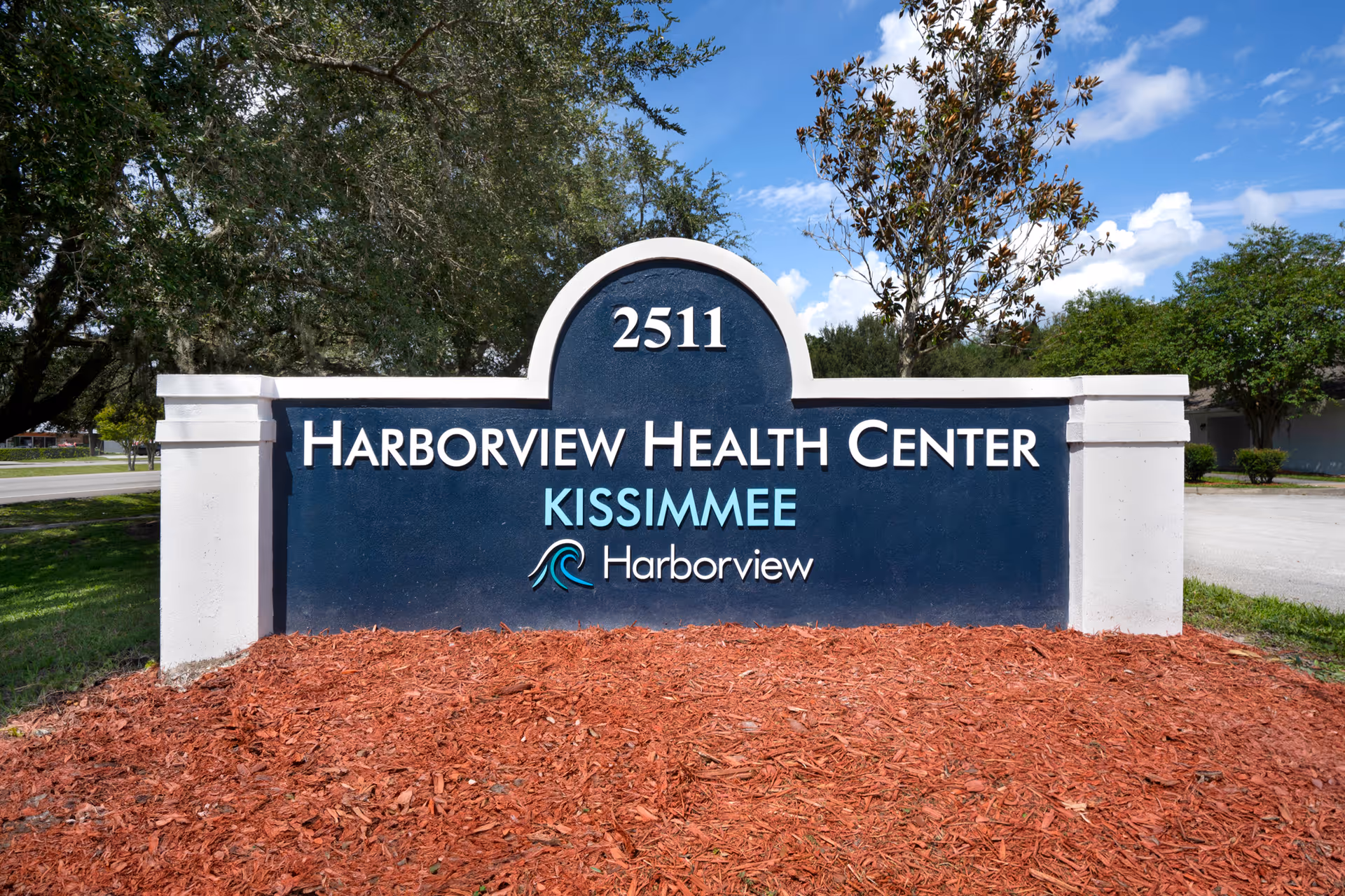 Outdoor sign for Harborview Health Center Kissimmee with the address number 2511, surrounded by trees and mulch under a blue sky with some clouds.