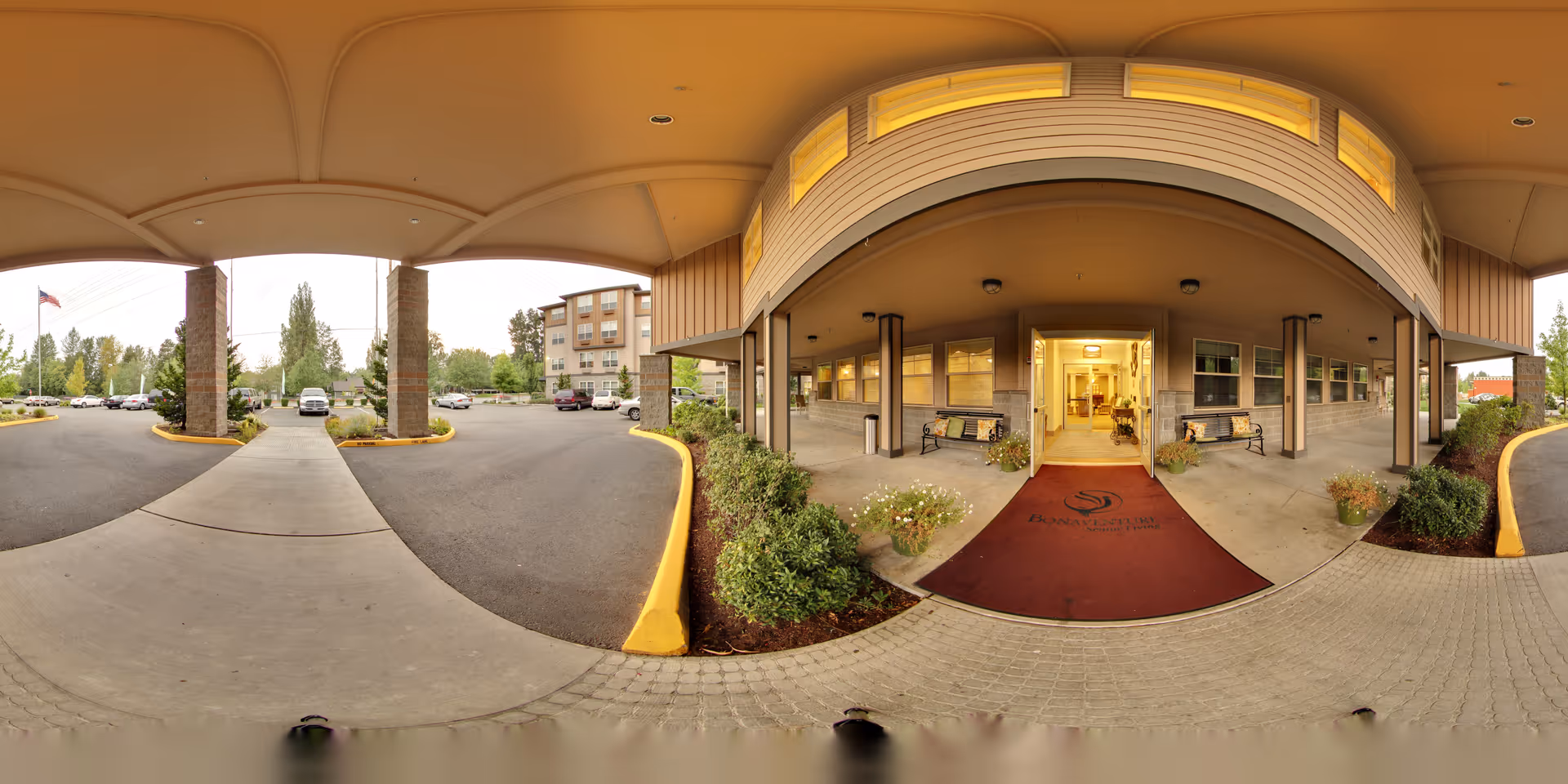 Covered entrance area of North Creek by Bonaventure senior living facility with a maroon welcome mat, benches on either side, potted plants, and a view of the parking lot and building exterior under an overhang.