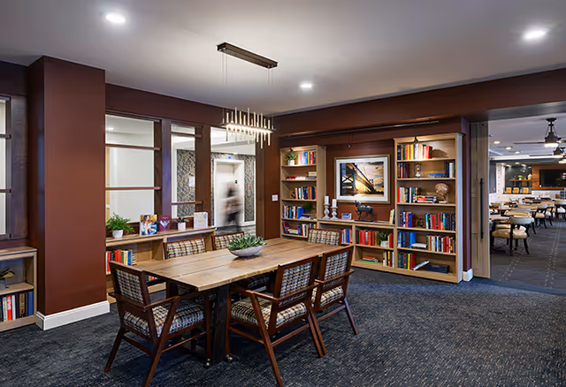 A cozy interior room with a wooden table surrounded by six cushioned chairs. The room features built-in bookshelves filled with books and decorative items, a modern chandelier hanging above the table, and a carpeted floor. In the background, there is an open doorway leading to another room with more seating and tables.