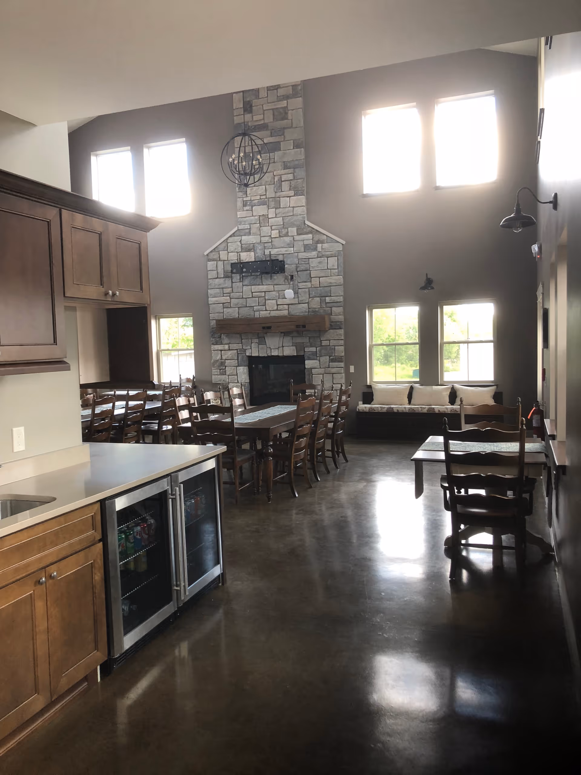 Interior view of a senior living facility dining and common area featuring a stone fireplace, multiple wooden dining tables with chairs, large windows letting in natural light, a kitchen counter with cabinets and a beverage cooler, and a cushioned bench under the windows.