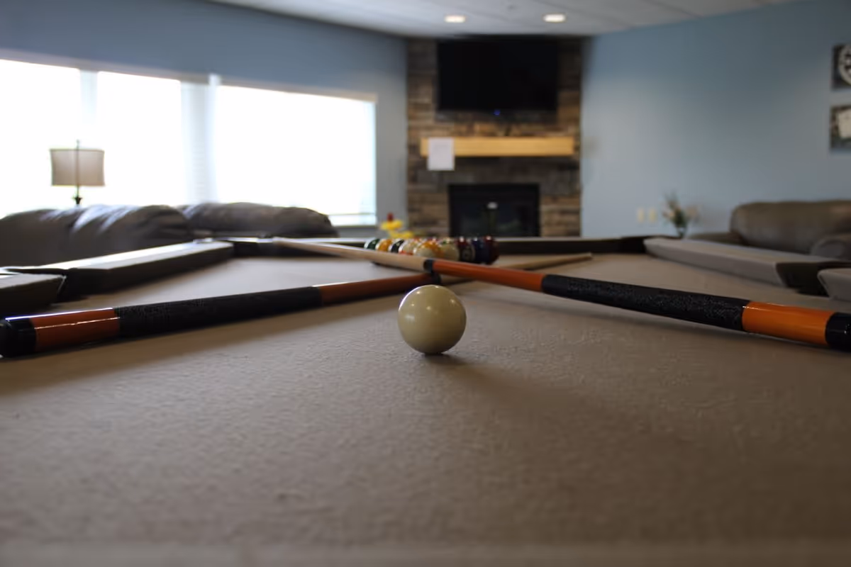 Close-up view of a pool table with two crossed pool cues and a white cue ball in the foreground. In the background, there is a stone fireplace with a mounted TV above it, a large window with blinds, and comfortable seating including sofas and a lamp.