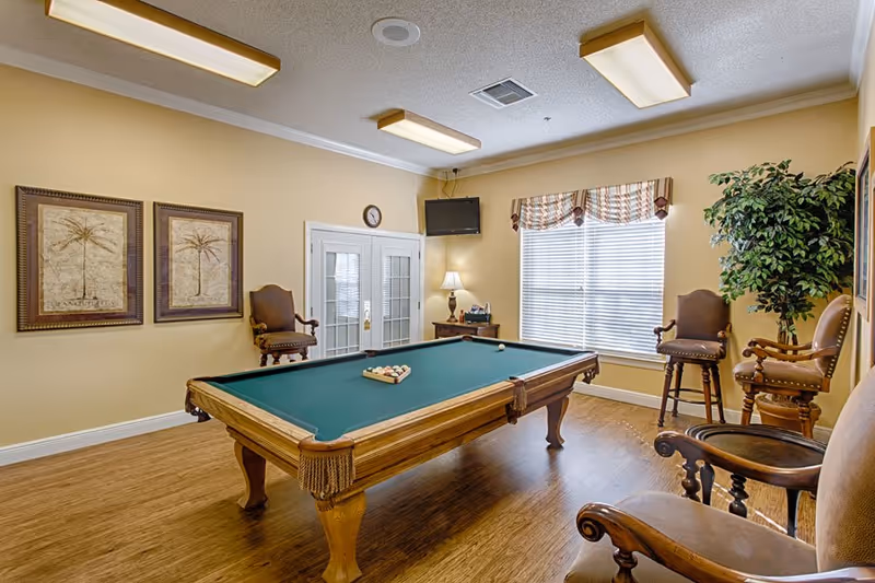 A well-lit room with a pool table in the center, surrounded by wooden chairs with cushioned seats. The room has beige walls, two framed palm tree artworks, a window with blinds and a valance, a small table with a lamp, a wall-mounted TV, and a potted plant in the corner.