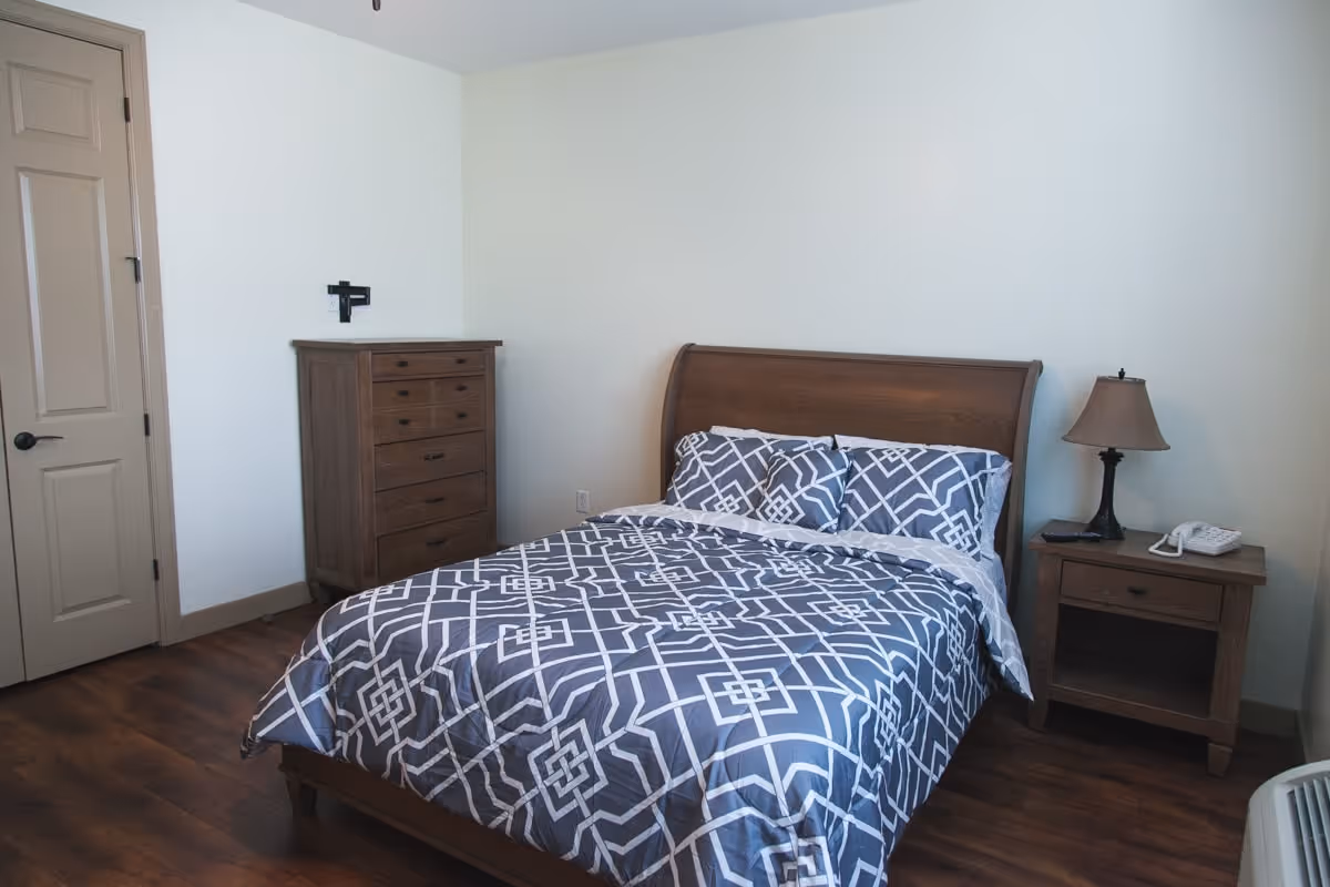 A bedroom with a wooden bed frame and a bed made with a blue and white geometric patterned comforter and matching pillows. To the right of the bed is a wooden nightstand with a lamp and a telephone. To the left is a wooden dresser with multiple drawers. The room has wooden flooring and light-colored walls.