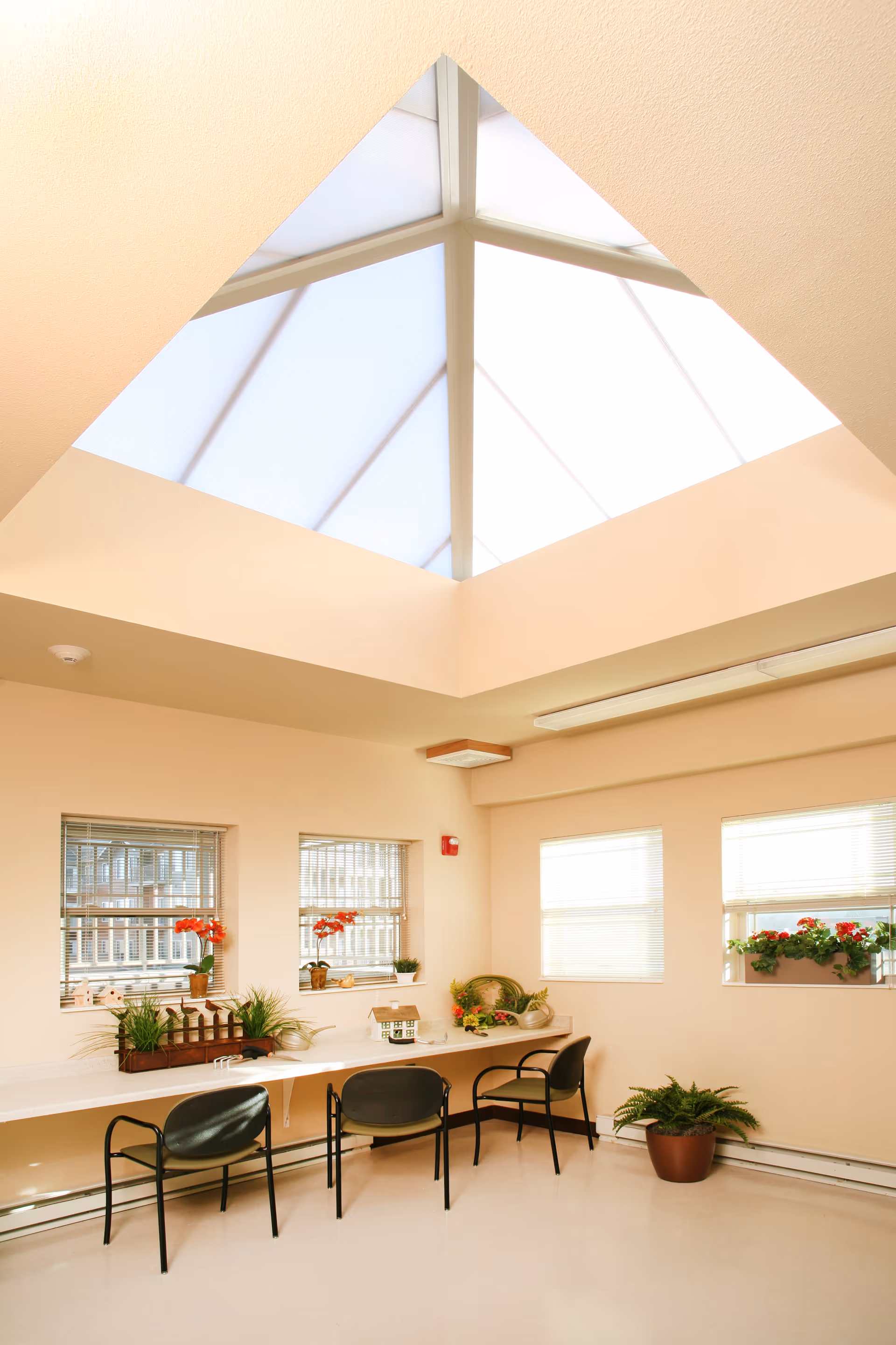 A bright room with a large skylight ceiling, three black chairs placed along a long counter under windows with blinds. The counter is decorated with small potted plants and flowers. The walls are light beige, and there is a potted plant on the floor near the corner.