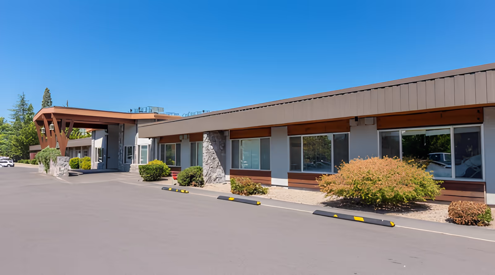 Exterior view of Hearthstone Nursing & Rehabilitation Center showing a single-story building with large windows, brown roof trim, stone pillars, and landscaped bushes along the front. The sky is clear and blue.