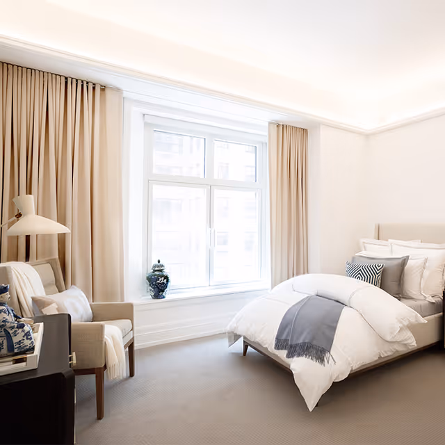 A bright and airy bedroom with a large window covered by beige curtains. The room features a neatly made bed with white bedding and gray accent pillows and throw. There is a beige armchair with a pillow and a white throw blanket next to a black side table with decorative items. The walls and carpet are light-colored, creating a clean and cozy atmosphere.