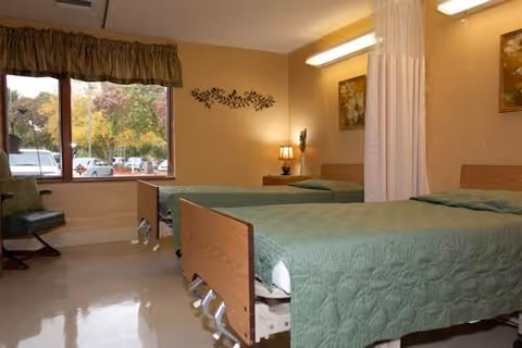 A rehabilitation or healthcare center room with two hospital beds covered in green quilts, a window with a view of trees and parked cars, a green chair, a nightstand with a lamp, wall decorations, and a privacy curtain between the beds.