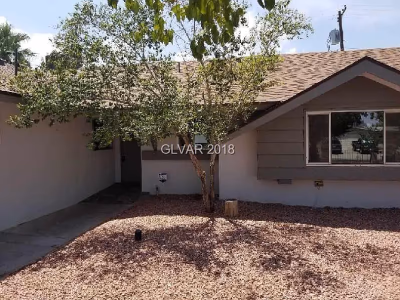 Front exterior view of a single-story house with a tree in the front yard and a gravel-covered ground. The house has a window on the right side and a covered entrance on the left.