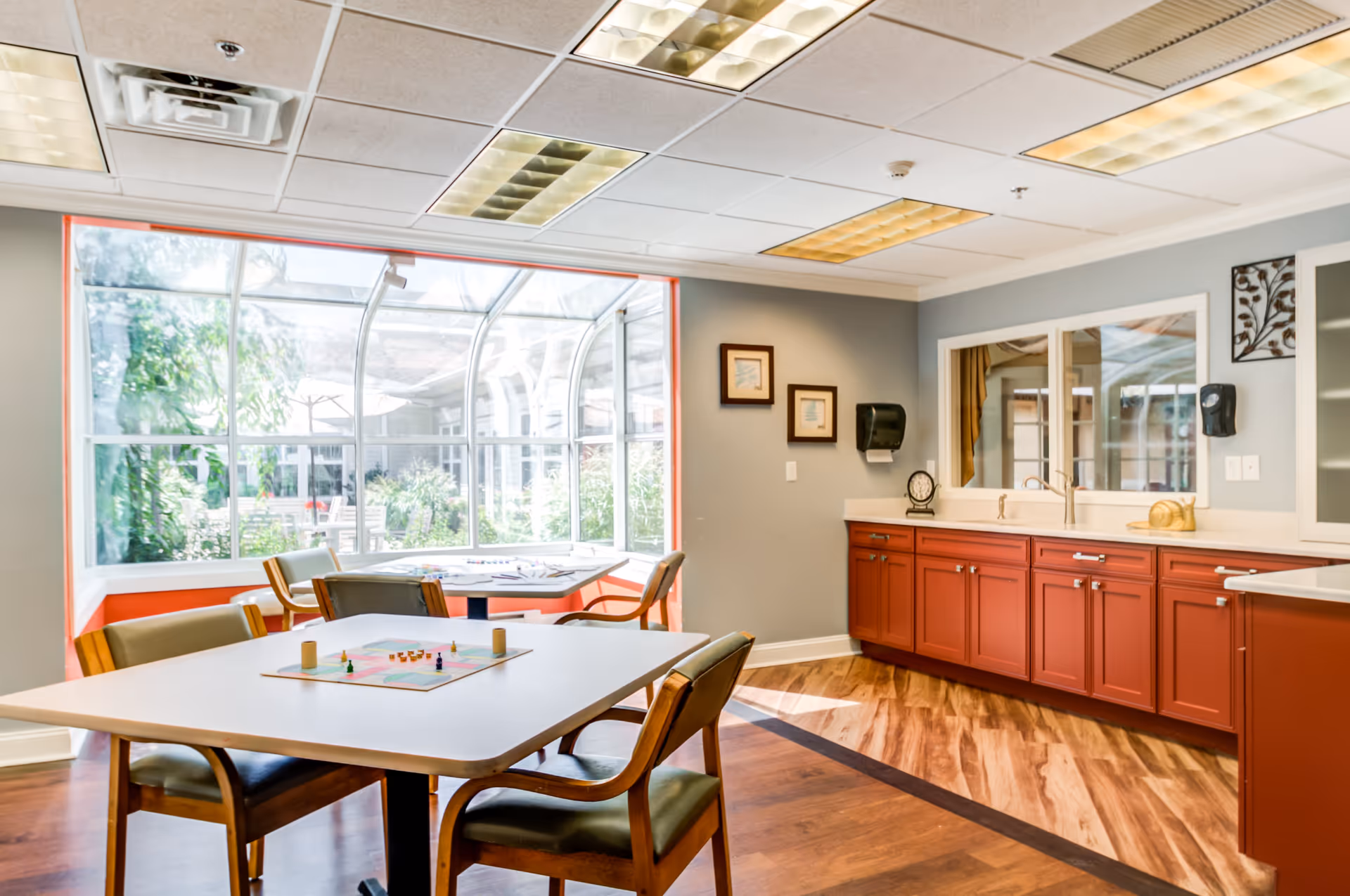 A bright and inviting common area with a large window overlooking a garden. The room features a table with chairs around it, a board game set up on the table, and red cabinetry with a countertop and sink along one wall. The floor has a wood pattern, and the walls are painted gray with framed artwork and a wall-mounted soap dispenser.