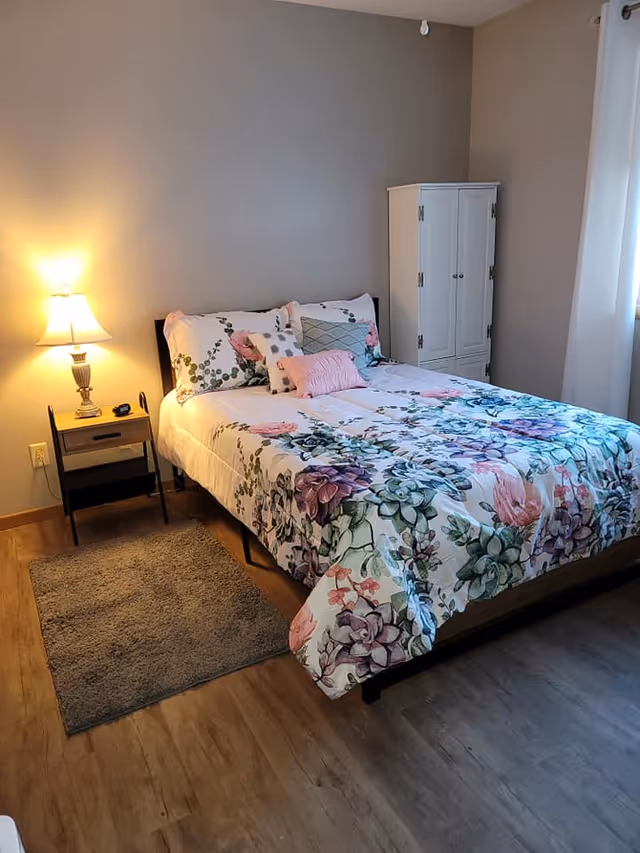 A cozy bedroom with a bed featuring floral bedding and multiple pillows. Next to the bed is a small wooden nightstand with a lamp and a small item on it. There is a gray rug on the wooden floor beside the bed. A white cabinet stands against the wall near a window with white curtains.