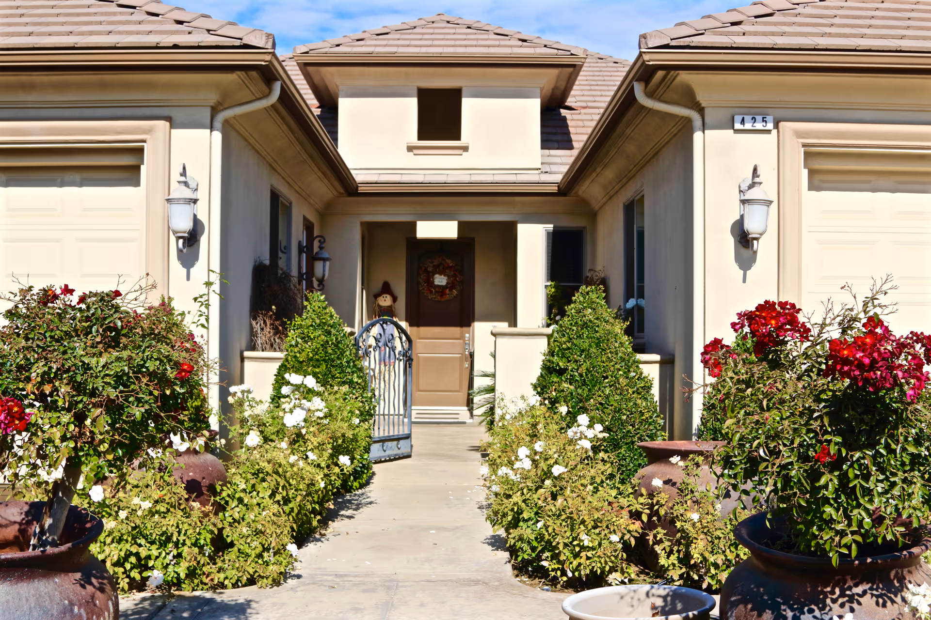 Front entrance of a residential building with a pathway lined by green bushes and flowering plants in large pots. The building has a beige exterior with two garage doors on either side of the entrance. A brown door with a wreath and a scarecrow decoration is visible at the end of the pathway. The house number 425 is displayed on the right side above the garage door.