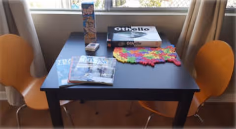 A small black table with two orange chairs on either side, placed in front of a window with beige curtains. On the table, there is a colorful puzzle in progress, a box of the game Othello, a box of crackers, a deck of cards, and two magazines.