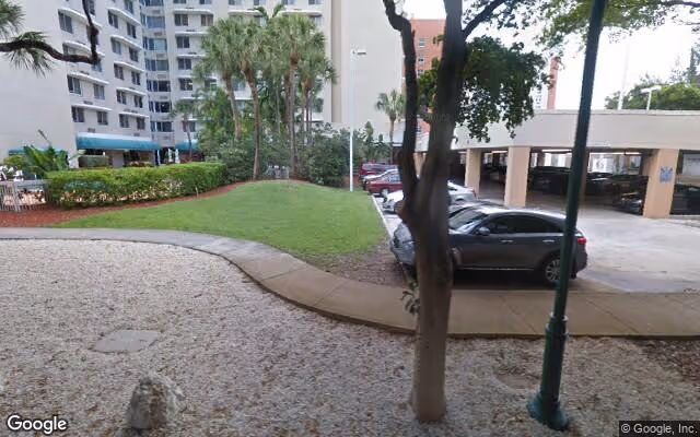 Outdoor view of a senior living facility showing a landscaped area with grass, trees, and a gravel section. There is a paved walkway curving through the area, parked cars, and a multi-story building in the background.